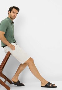 Outfit da uomo composto da una polo verde, pantaloni corti beige con tasche e sandali neri senza lacci, con una sedia di legno come accessorio.