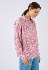 Camisa de manga larga con botones en rosa pálido con un patrón floral que presenta flores rojas, blancas y amarillas, confeccionada con una tela ligera.