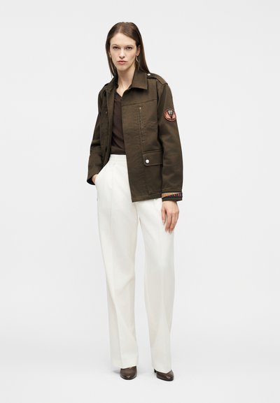 Γυναίκα που φοράει χακί πράσινο μπουφάν με μπαλώματα, καφέ τοπ, λευκό παντελόνι με φαρδιά πόδια και καφέ μυτερά παπούτσια, στέκεται με τα χέρια στις τσέπες.