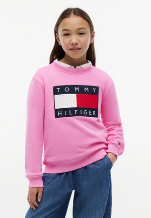 Κορίτσι που φοράει ένα ροζ φούτερ Tommy Hilfiger και μπλε τζιν, στέκεται με το ένα χέρι στην τσέπη μπροστά σε λευκό φόντο.