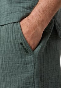 Shorts en tissu texturé vert avec une poche visible et un subtil détail brodé "SEASIDE" sur le côté.