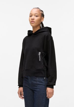 Black cropped hoodie made of cotton blend, featuring a drawstring, graphic text on the front, and ribbed cuffs. Paired with dark jeans.
