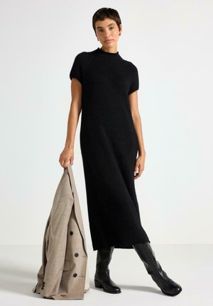 Vestido negro de punto suave, de manga corta y silueta recta. Modelo sostiene una chaqueta ligera gris claro y lleva botas negras hasta la rodilla.