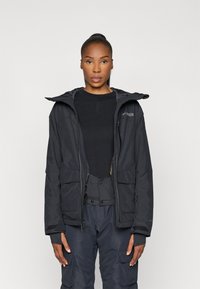 Chaqueta impermeable negra con capucha, que presenta dos bolsillos frontales, cierre con cremallera y puños ajustables. Lleva puesta sobre una capa base negra.