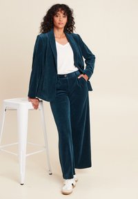 Completo in velluto a coste teal con pantaloni a gamba larga e blazer su misura. Indossato sopra una maglietta bianca a scollo a V, completato da sneakers bianche.