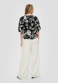 Zwarte blouse met grote witte bloemenprint, korte mouwen en een losse pasvorm, gecombineerd met witte wijde broekspijpen. Opvallende gladde textuur.