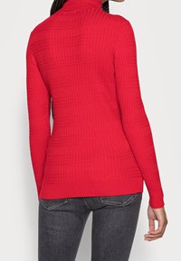Red, ribbed turtleneck sweater with textured pattern, long sleeves, and a fitted design. Paired with dark jeans.