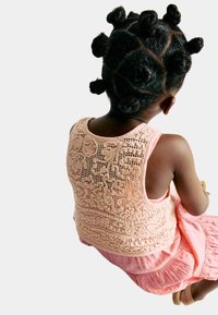 Haut sans manches rose clair avec des détails en dentelle élaborés dans le dos, associé à une jupe rose à volants. Cheveux coiffés en plusieurs chignons arrondis.