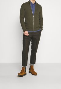 Man som bär en olivgrön zip-up strukturtröja, blå pikétröja, mörka avklippta byxor, grå strumpor och tan mocka Chelsea boots står.