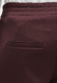 Bordeauxfarvede bukser med en struktureret finish, elastisk talje og en enkelt baglomme med synlig syning. Glat stof, skræddersyet design.