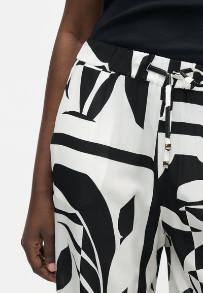 Άτομο που φοράει μαύρο και άσπρο παντελόνι με αφηρημένο σχέδιο και κορδόνι στη μέση, με μέρος ενός μαύρου τοπ ορατό.