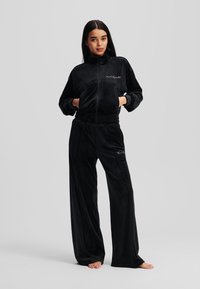 Melns vilnas treniņtērps ar rāvējslēdzēja jakas un elastīgām biksēm. Iezīmes sānu kabatas un smalks zīmols uz krūškurvja un augšstilba. Gluda tekstūra.