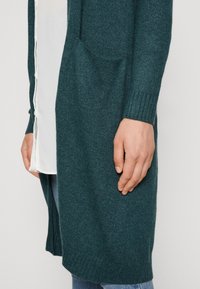 Tirkīza garā cardigan, izgatavots no adītas materiāla, ar divām kabatām, ribu aprocēm un atvērtu priekšu. Uzvilkts virs baltā krekla.