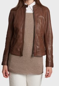 Hnědá kožená bunda s přiléhavým střihem, s kapsami na zip a stojáčkovým límcem, vrstvená přes béžový pletený svetr. Minimalistický styl.