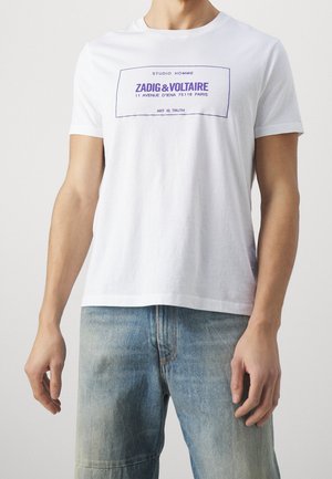 Mann trägt ein weißes T-Shirt mit kurzen Ärmeln und dunkelblauem Schriftzug "Zadig & Voltaire" sowie ausgewaschene blaue Jeans, steht vor neutralem Hintergrund.