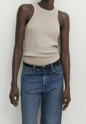 Personne à la peau foncée portant un haut beige sans manches côtelé rentré dans un jean bleu taille haute avec une ceinture noire, debout contre un mur uni.