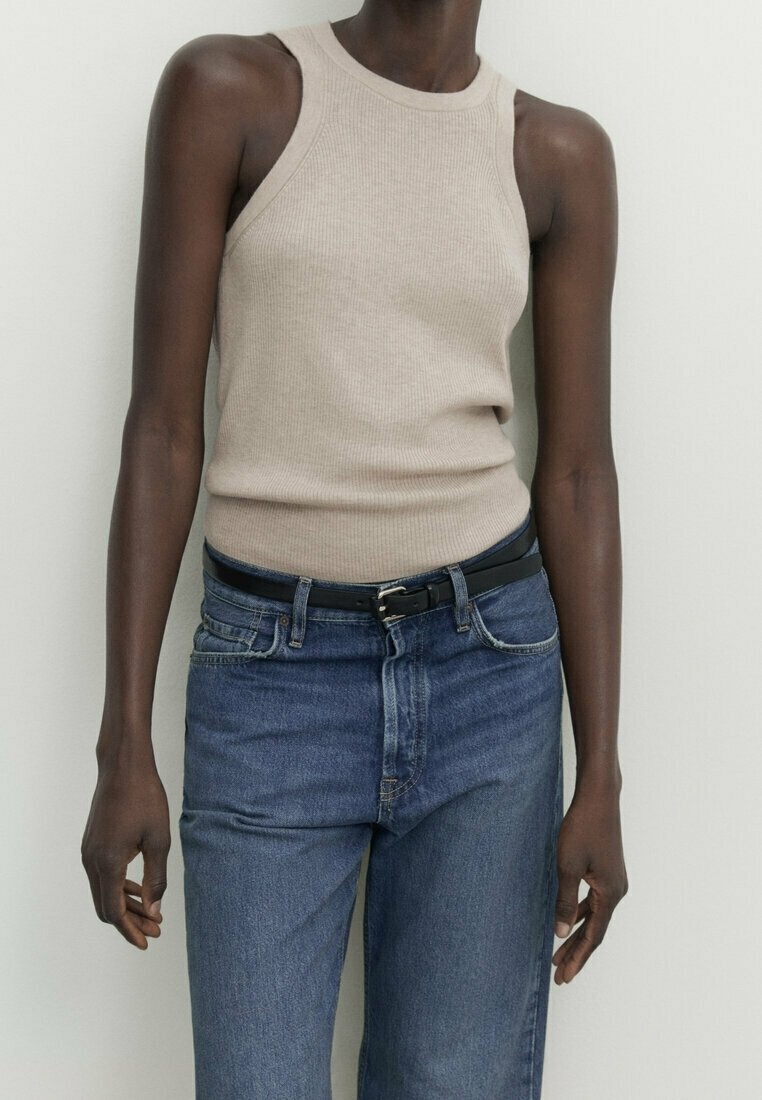 Personne à la peau foncée portant un haut beige sans manches côtelé rentré dans un jean bleu taille haute avec une ceinture noire, debout contre un mur uni.