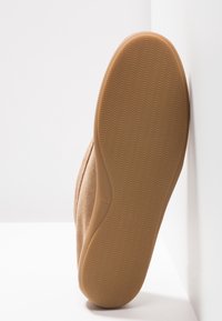 Semelle d'une chaussure en daim beige avec un motif ondulé, positionnée à la verticale contre un mur blanc et un sol blanc.
