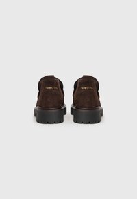 Chaussures en daim marron avec une semelle en caoutchouc noir, dotées d'un talon rond et d'une languette en boucle à l'arrière pour un enfilage facile. Logo de la marque doré sur le talon.