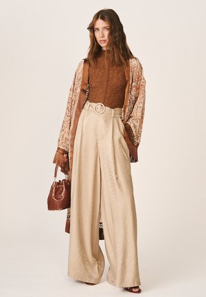 Femme portant un pantalon beige large avec ceinture, un haut marron texturé, un kimono à motifs, tenant un sac à main marron texturé sur fond uni.
