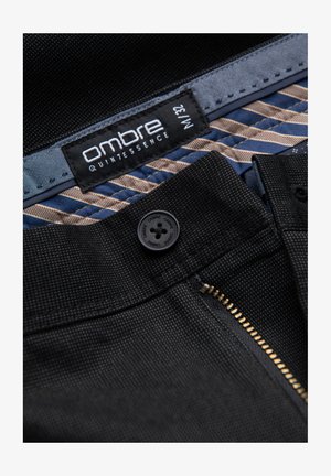 Tejido texturizado negro con un cierre de cremallera dorado, cierre de botón negro y una etiqueta que dice "quintessencia ombré" en un forro azul con patrón.