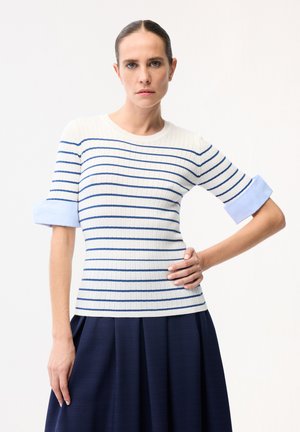 Kobieta z włosami związanymi do tyłu, ubrana w biały sweter z niebieskimi poziomymi paskami i jasnoniebieskimi rękawami podwiniętymi, w połączeniu z granatową spódnicą.