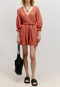 Koraalrode wikkeljurk van gestructureerde stof met volumineuze mouwen, aangekleed met een strakke taille, gecombineerd met zwarte flat sandals en een zwarte handtas.