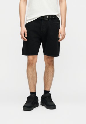 Personne portant un short en denim noir avec une ceinture noire, des baskets noires, des chaussettes noires et un t-shirt blanc sur un fond uni.