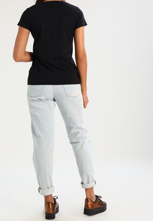 Femme portant un t-shirt noir, un jean bleu clair retroussé avec un patch, et des chaussures lacées en bronze métallisé, debout, dos tourné.