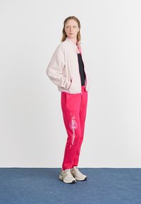 Růžová bomber bunda s kontrastními růžovými lemy, spárovaná s růžovými tepláky se bílým textem. Model má na sobě černý top a béžové boty.
