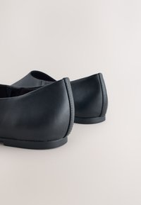 Paire de chaussures plates en cuir noir sans lacets, vues de dos sur une surface claire unie.