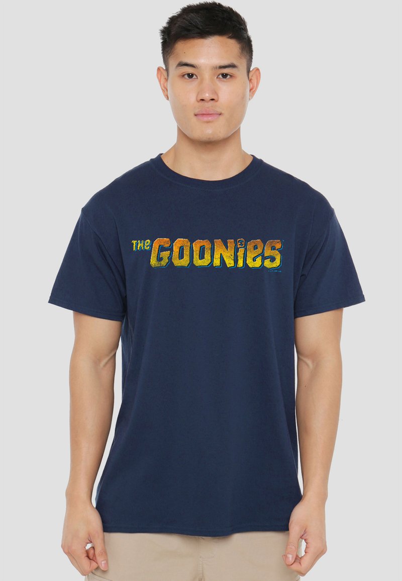 Young man wearing a navy blue T-shirt with yellow and orange "The Goonies" text, standing against a plain light gray background.