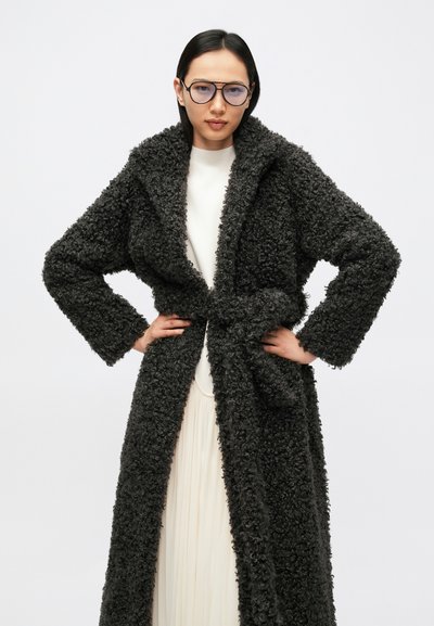 Tamnosivi plišani kaput s teksturiranim, kovrčavim materijalom, s pojasom za vezanje i širokim reverima, nošen preko svijetle haljine.