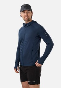 Homme portant un sweat à capuche bleu marine zippé avec des passe-pouces, un short noir et une casquette noire, regardant sur le côté avec un léger sourire.