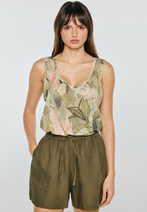 Młoda kobieta ubrana w bezrękawnik z botanicznym nadrukiem, wpuszczony w krótkie spodenki z wysokim stanem w kolorze oliwkowej zieleni, z rękami w kieszeniach.