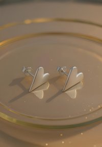 Orecchini a perno a forma di cuore in argento con superfici lisce e opache e bordi lucidi, che riflettono la luce su uno sfondo morbido e dorato.