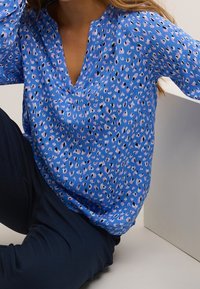 Blå bluse med V-halsmønster, der har lyserøde og sorte pletter. Blød stof med en afslappet pasform, parret med mørke bukser.