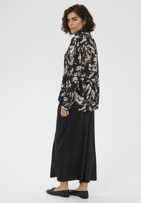 Juoda gėlėtu raštu suknelė ilgomis rankovėmis, derinama su ilgu juodu sijonu. Modelis dėvi juodus butus be kulnų ir stovi profilio pozicijoje.