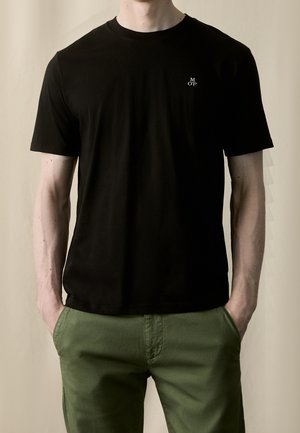 Czarny bawełniany t-shirt z krótkimi rękawami, okrągłym dekoltem i małym białym logo na lewej piersi, noszony z zielonymi spodniami.