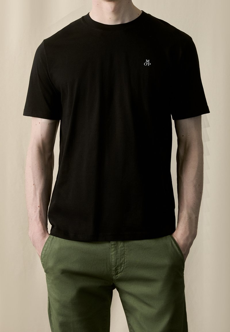 T-shirt en coton noir à manches courtes, avec un col rond et un petit logo blanc sur la poitrine gauche, porté avec un pantalon vert.