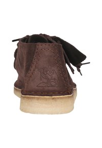 Brun mockaskor med en baksidestämpel som visar en kamel och en person. Mjuk textur, platt sula med kontrasterande ljusbrun sömnad.