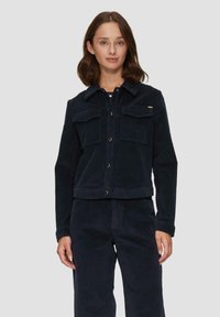 Granatowa kurtka z korduroyu z zapięciem na guziki, dwoma kieszeniami na piersi oraz dopasowanymi spodniami. Posiada wyraźny kołnierz i subtelne detale marki.