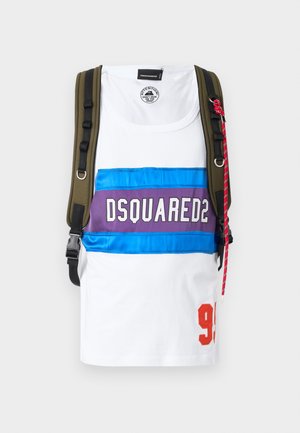 Jeune homme debout, portant un débardeur blanc avec le logo "DSQUARED2", un jean sombre déchiré, des baskets blanches et un sac à dos sur fond uni.