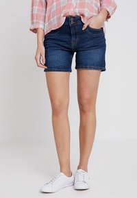 Tummansinisiä farkkushortsit korkealla vyötäröllä, joissa on napin kiinnitys, etutaskut ja käänteet. Yhdistettynä valkoisiin tennareihin.