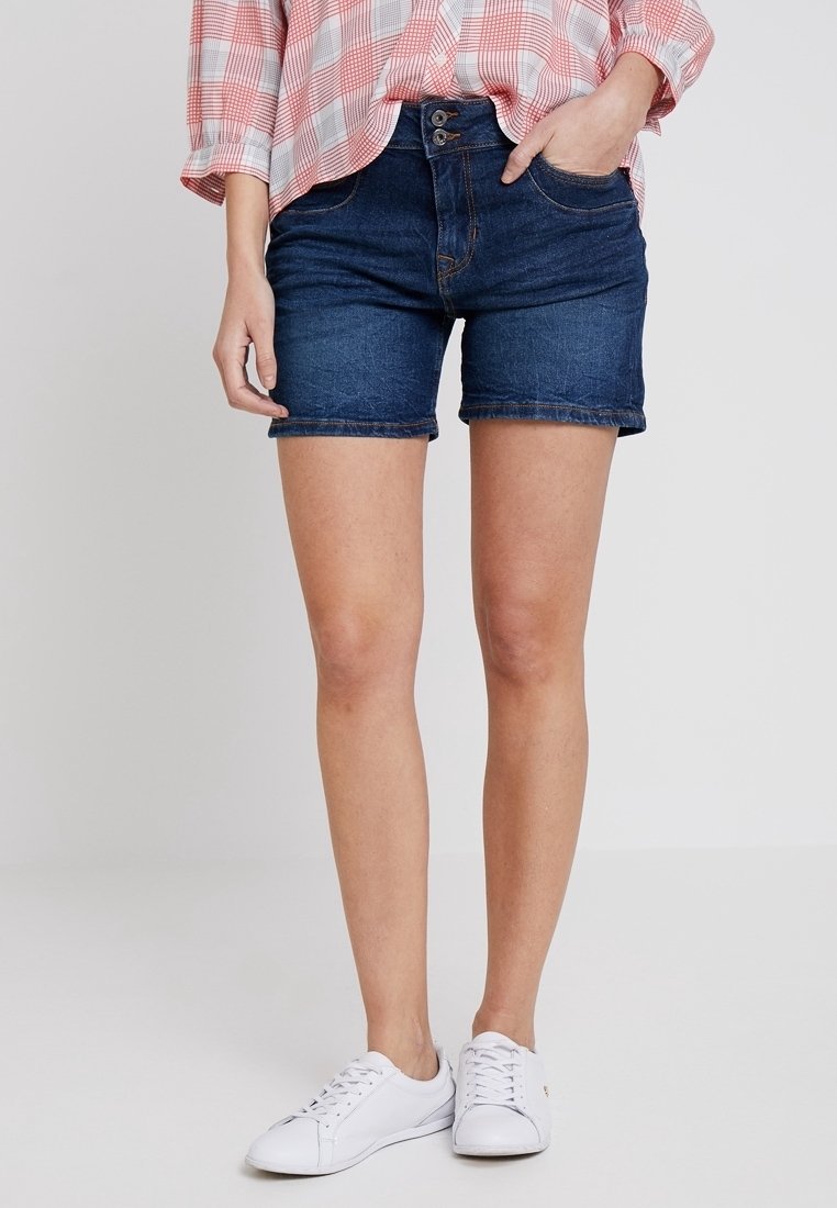 Tummansinisiä farkkushortsit korkealla vyötäröllä, joissa on napin kiinnitys, etutaskut ja käänteet. Yhdistettynä valkoisiin tennareihin.