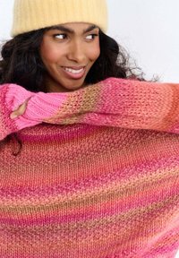 Kobieta z kręconymi włosami, ubrana w żółtą czapkę typu beanie oraz wielokolorowy, różowo-pomarańczowy sweter na drutach, uśmiechająca się i patrząca na bok.