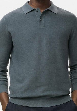 Mann trägt ein langärmliges, strukturiertes graues Poloshirt mit einem geknöpften Kragen und gerippten Bündchen, in eine dunkle Hose gesteckt.