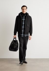 Μαύρο μονωμένο μπουφάν με κουκούλα, φορεμένο πάνω από πουκάμισο σε μαύρο και γκρι καρό. Σκούρα παντελόνια και μαύρα υποδήματα. Κρατάει μία μαύρη τσάντα duffel.