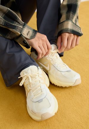 Ręce wiążące sznurowadła kremowych butów Nike, osoba ubrana w ciemne spodnie i zieloną kratkowaną kurtkę, siedząca na żółtej powierzchni.