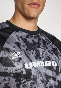 Grau und schwarzes Sporttrikot mit geometrischem Muster und weißem "UMBRO"-Logo. Verfügt über einen Rundhalsausschnitt und strukturierter Stoff.